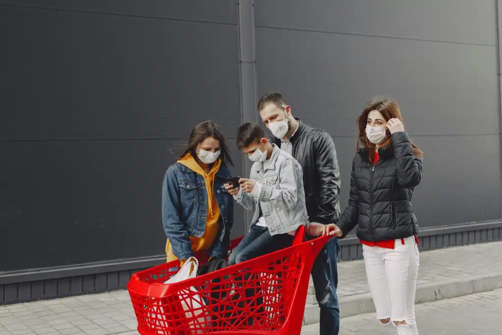 Happy family in casual clothes and medical masks standing on street after shopping while kid sitting in trolley and playing games on smartphone